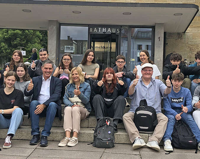 Schülerinnen und Schüler sitzen auf der Treppe vor dem Rathaus und zeigen den Daumen hoch